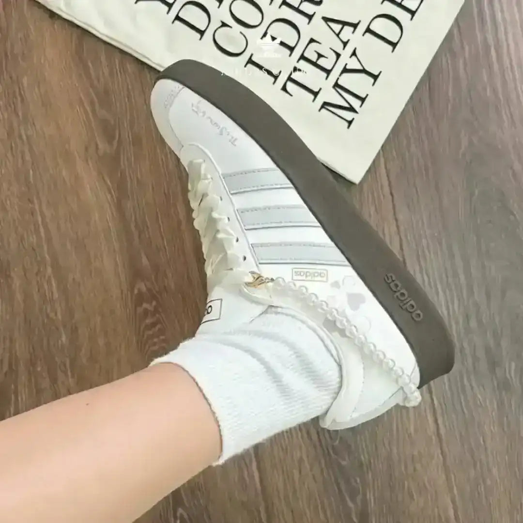 Adidas white leather sneakers accented with grey stripes and a pearl ankle bracelet, supported by a dark gum sole for a clean minimalist look.