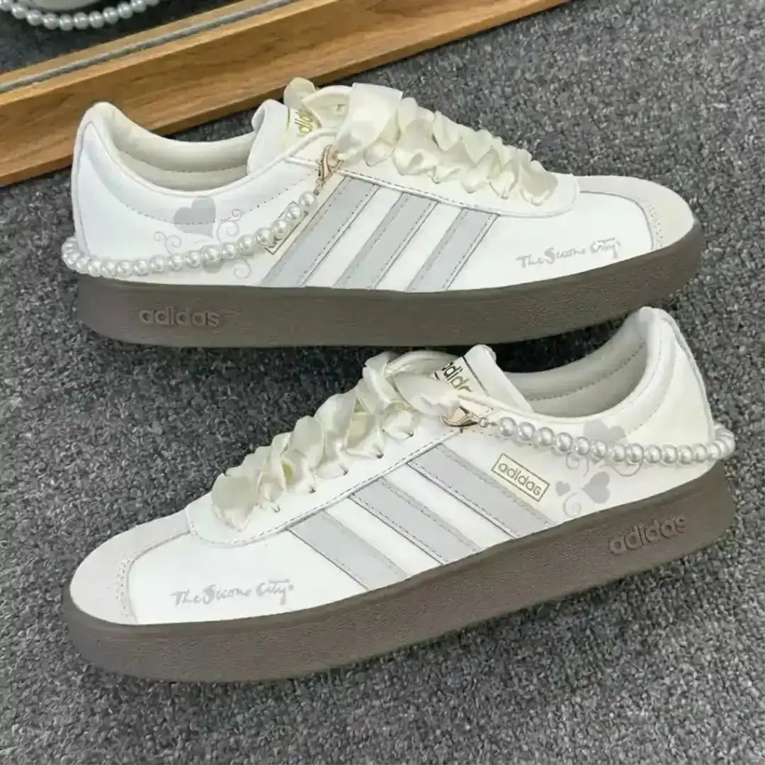 Adidas white sneakers highlighted by grey stripes and a pearl ankle chain, supported by a dark gum outsole for a refined lifestyle silhouette.