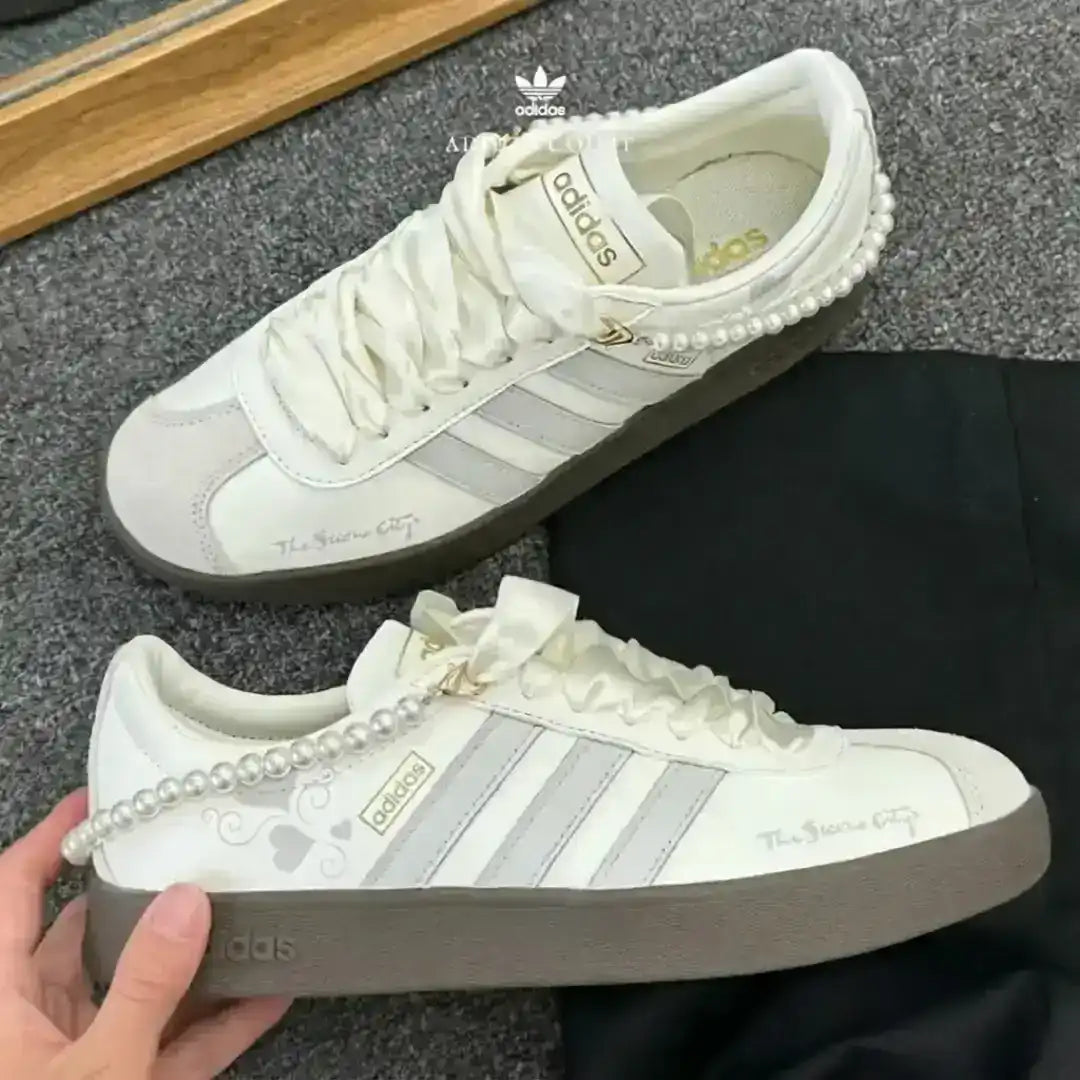 Adidas sneakers crafted in white leather with grey stripes and a pearl ankle detail, paired with a dark gum sole for casual sophistication.