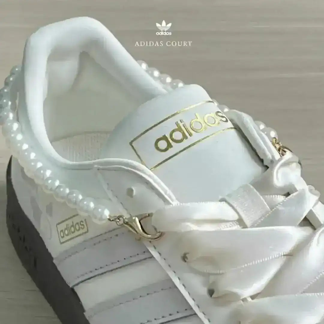 White Adidas shoes with grey triple stripes and a decorative pearl ankle bracelet, showcasing a dark gum sole for a trendy lifestyle look.
