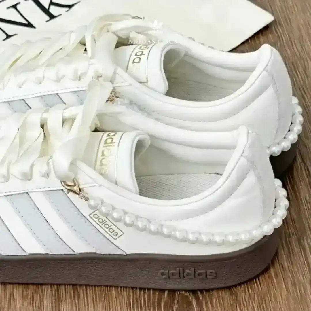 Adidas white sneakers with grey branding stripes and a pearl ankle bracelet, paired with a dark gum sole for a modern and elegant twist.