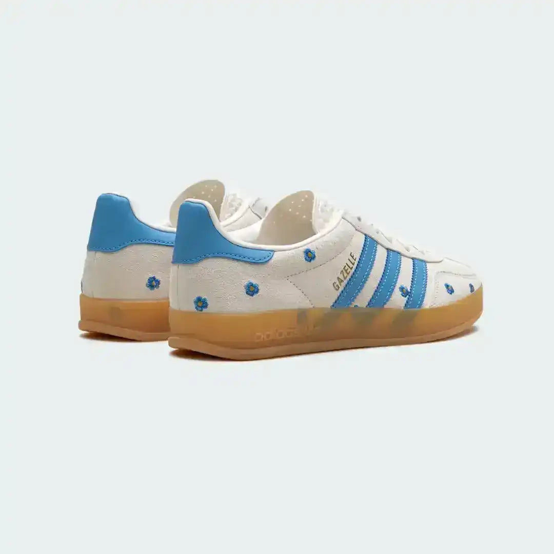 White Adidas Gazelle shoes featuring embroidered blue flowers, premium suede texture, iconic blue stripes and a vintage-style translucent gum sole.