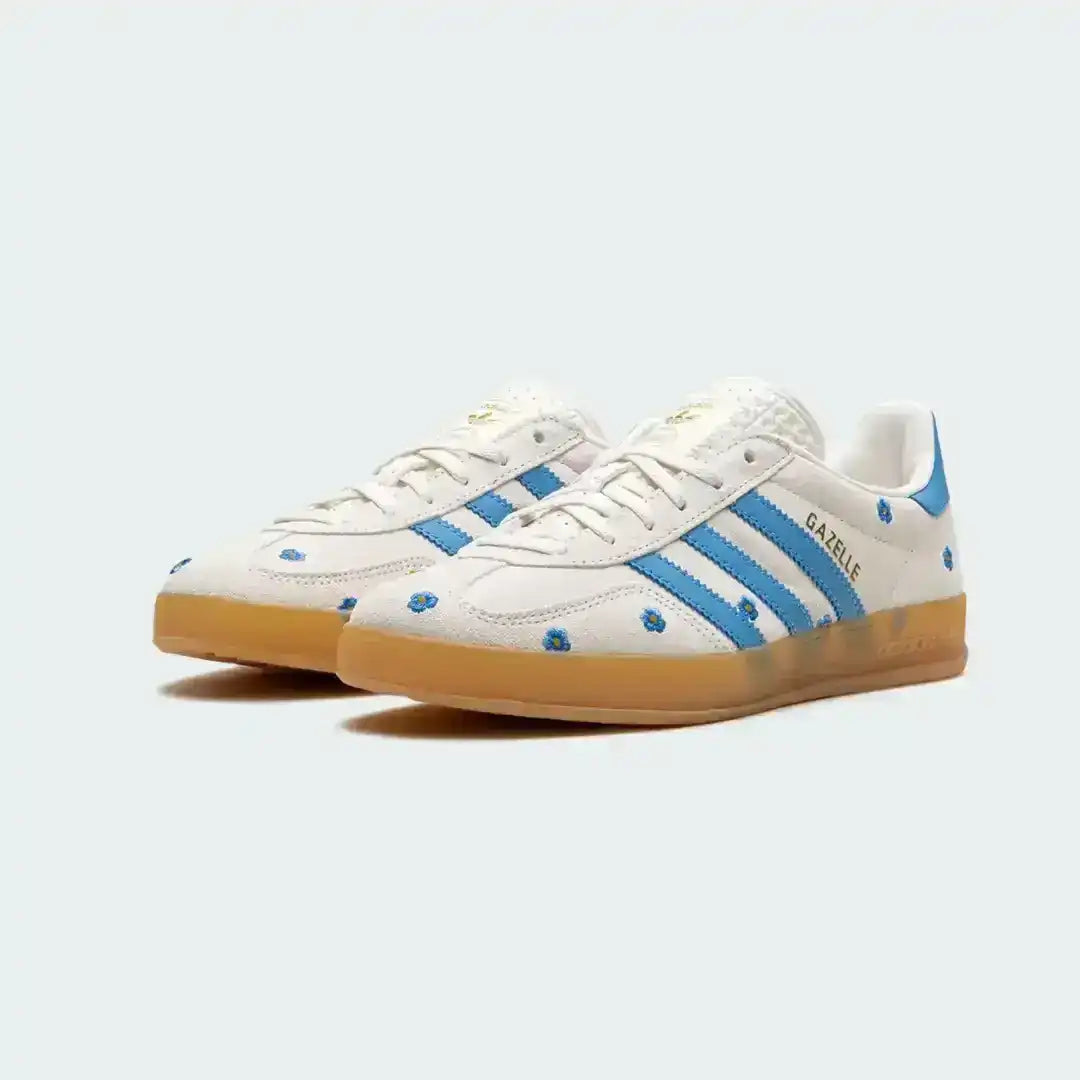 White Adidas Gazelle sneakers showcasing small blue embroidered flowers, iconic blue stripes, premium materials and a lightly translucent gum sole.