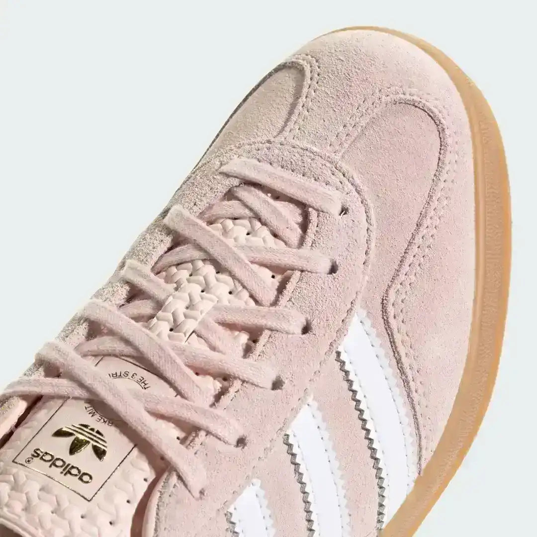 A pair of pink suede Adidas Gazelle Indoor shoes, showcasing the retro aesthetic with gold 'GAZELLE' text on the side and a textured geometric pattern on the tongue.