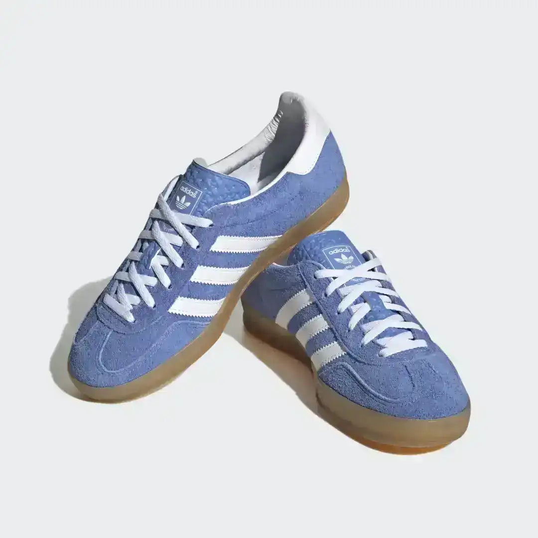 Adidas Gazelle Indoor sneakers featuring a premium blue suede upper, contrasting white serrated three stripes, and a signature translucent gum rubber outsole.