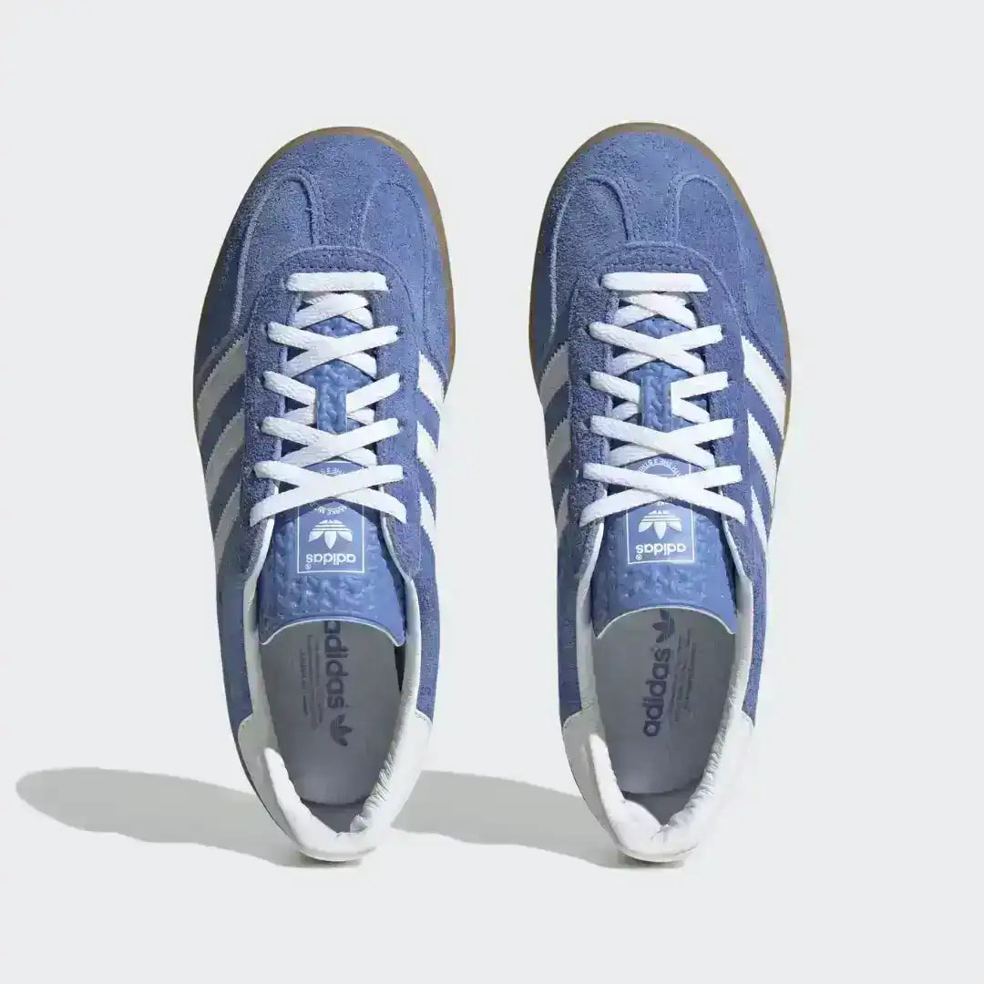 Adidas Gazelle Indoor footwear featuring a blue textured upper, white serrated stripes, and a distinct gum midsole that wraps up the sidewall.