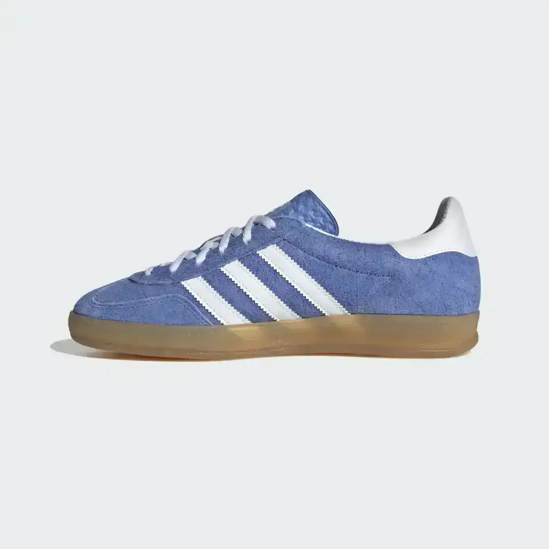 Adidas Gazelle Indoor sneakers featuring a premium blue suede upper, contrasting white serrated three stripes, and a signature translucent gum rubber outsole.