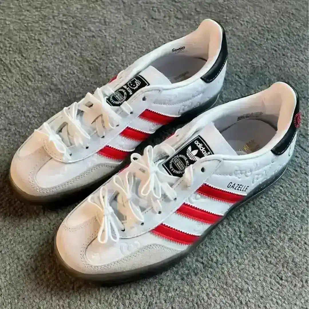 Side view of the Hello Kitty x Adidas Gazelle Indoor shoes in white with vivid red stripes, showcasing a black heel tab with bow detail, grey suede toe overlay, and a cute character face on the heel.