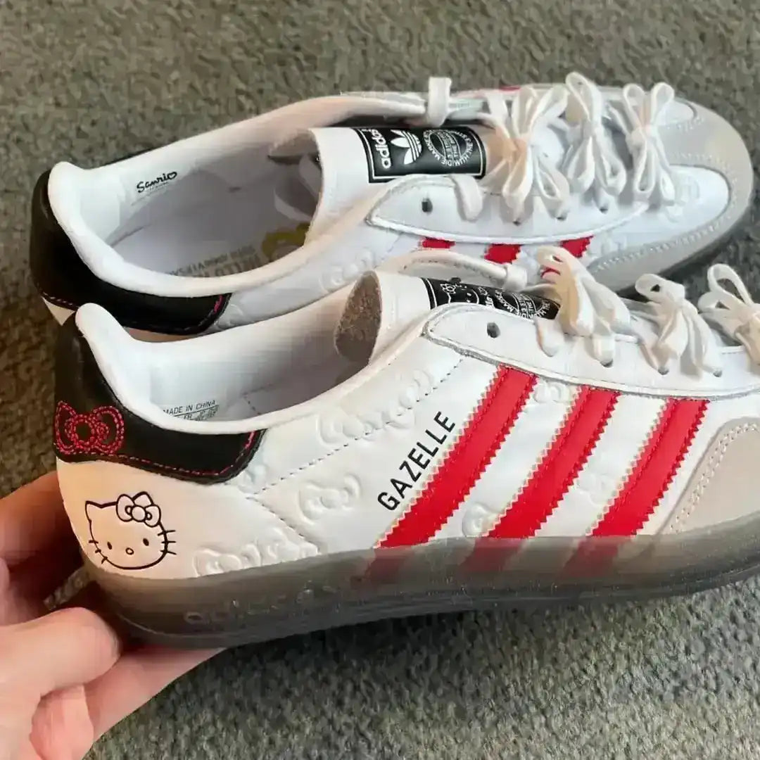 Exclusive Hello Kitty Adidas Gazelle Indoor sneakers in white and red colorway, highlighting the grey suede T-toe, red serrated stripes, and playful Hello Kitty face graphic printed on the lateral heel.