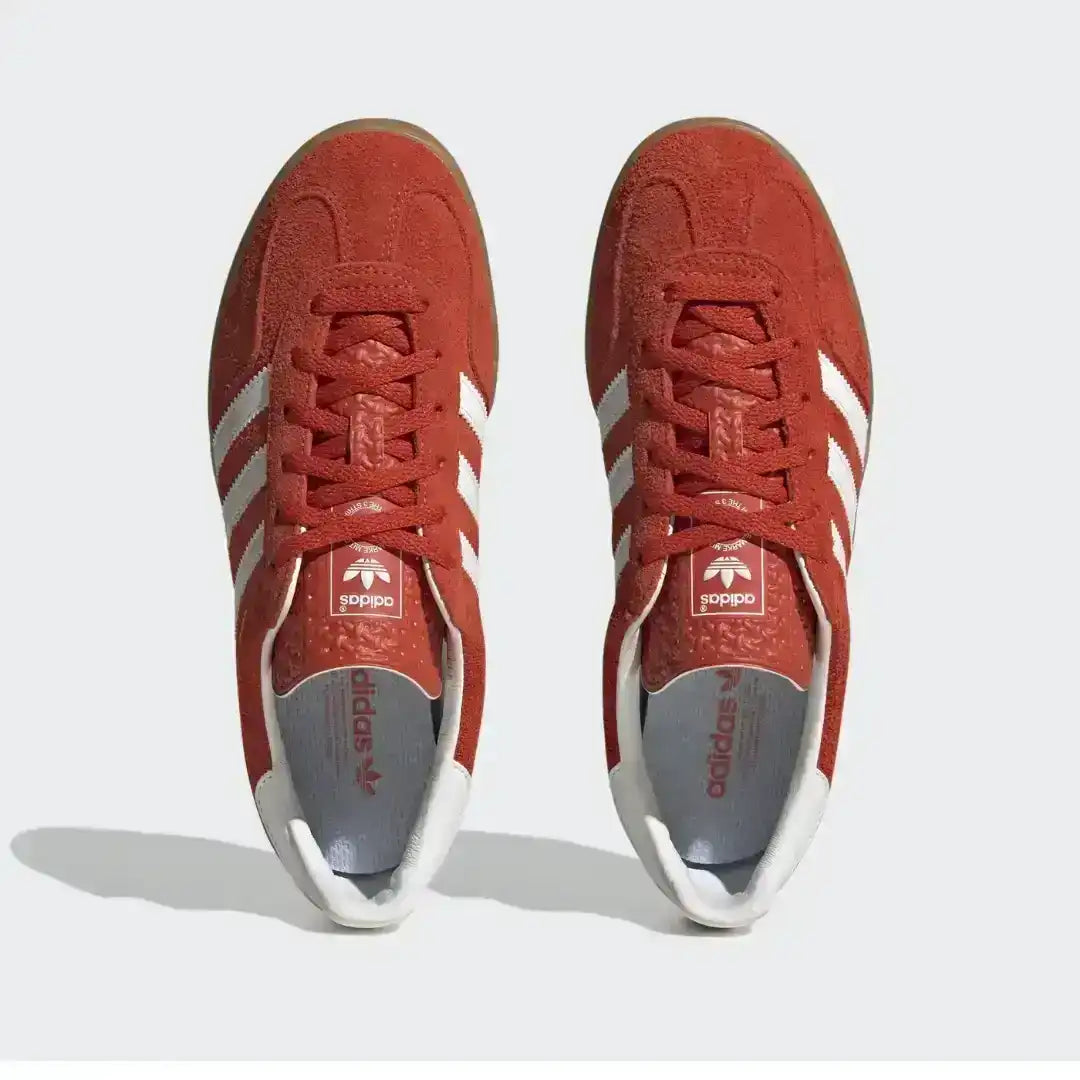 Adidas Gazelle Indoor shoes in a bold red suede colorway with white stripes. The design features a low-profile silhouette and a distinct dark gum midsole that wraps up the side.