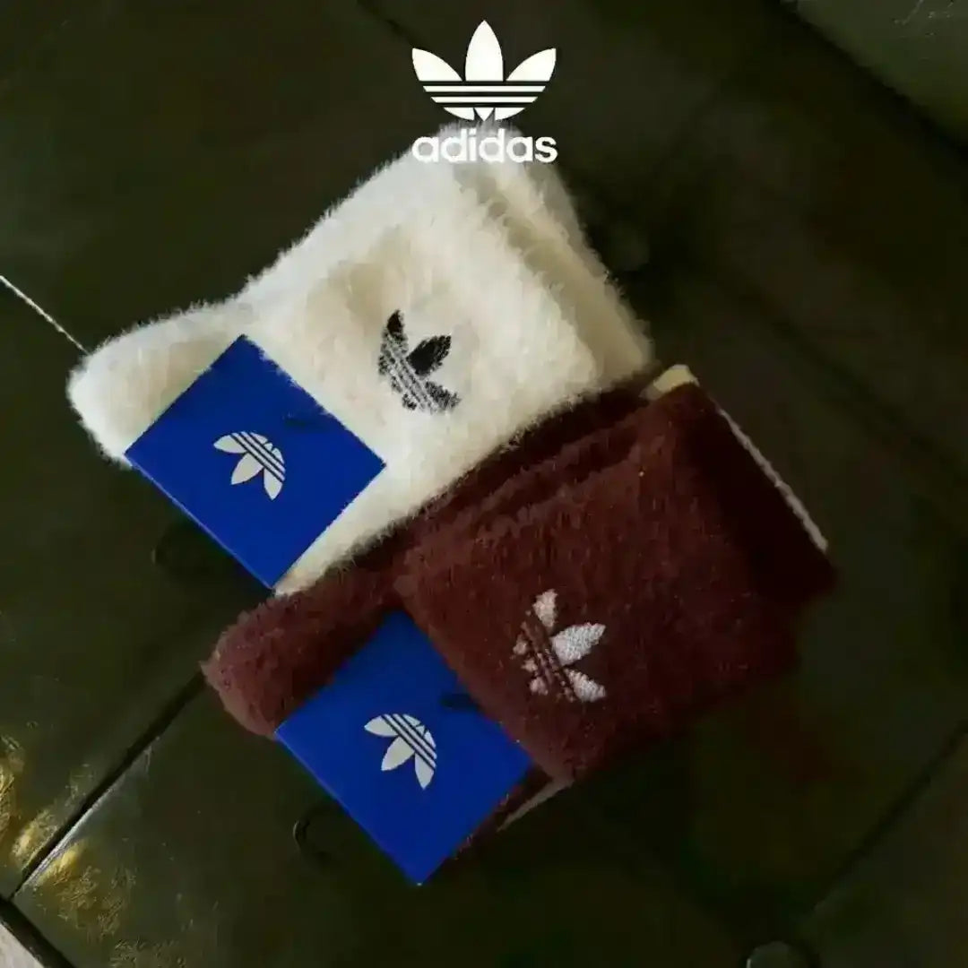 Adidas Originals fluffy socks displayed in black, white, and rust brown colors, all featuring a soft cozy texture and the classic embroidered Trefoil branding on the cuffs.