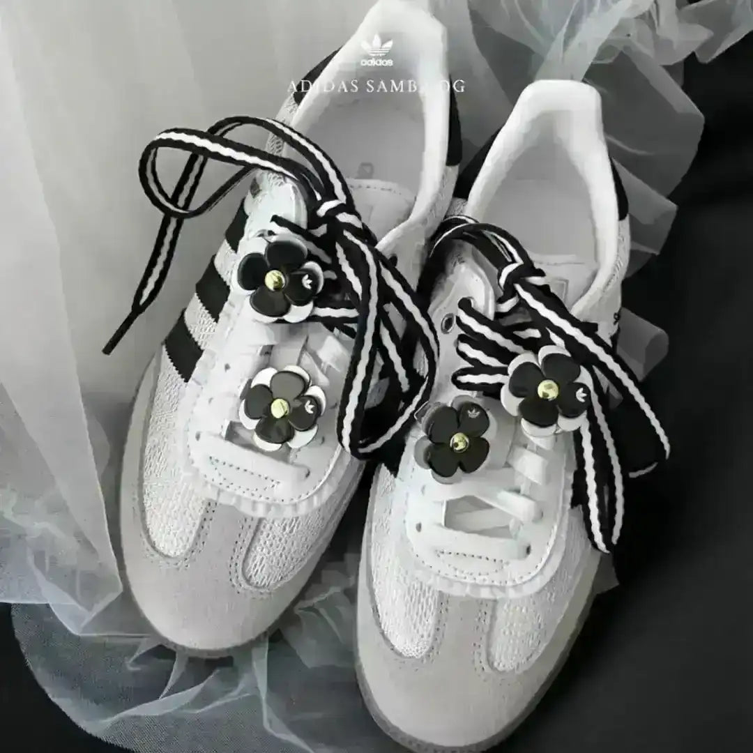 A pair of white Adidas Samba OG sneakers customized with thick black and white striped laces and black flower charms, presented on a soft fabric background.