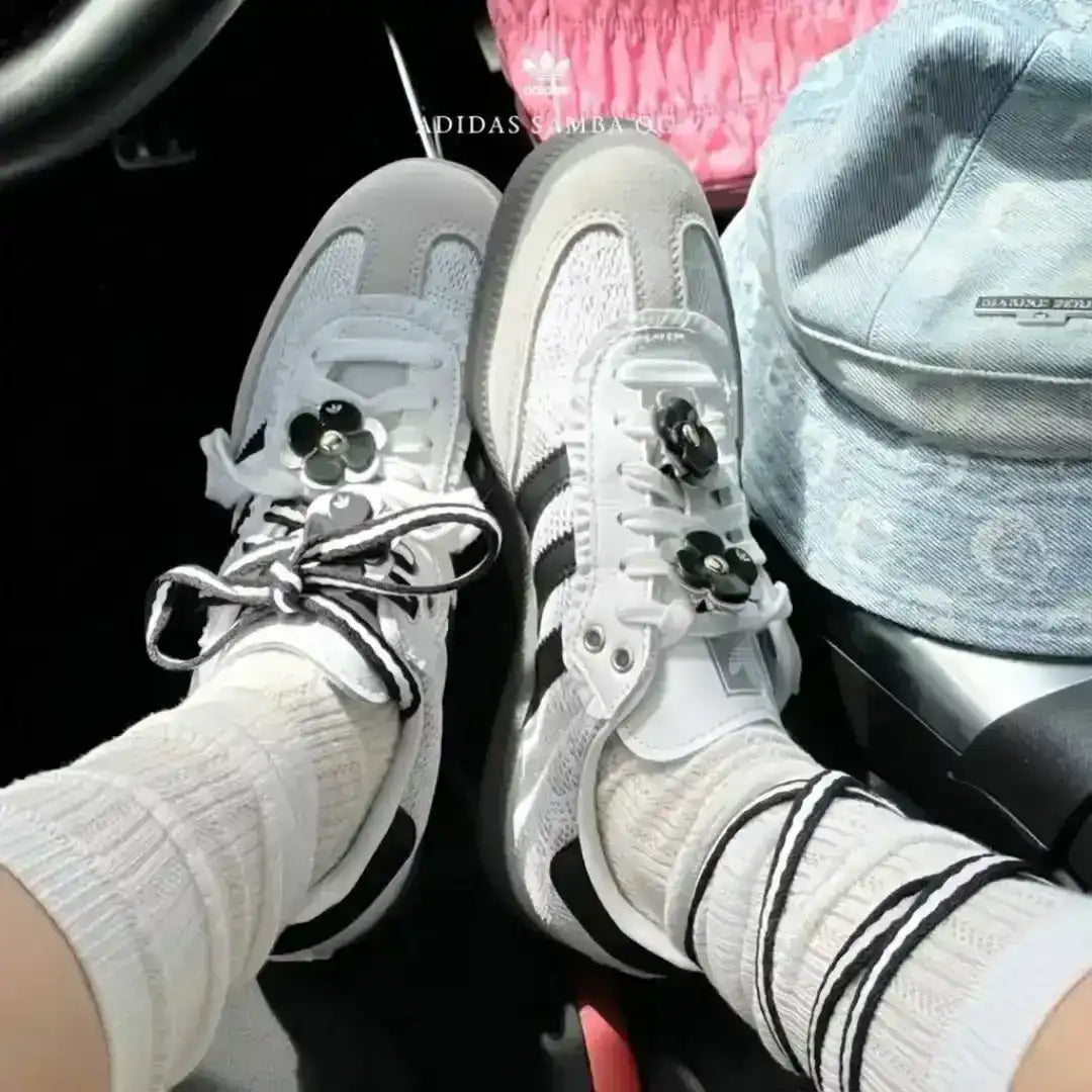 Custom Adidas Samba OG sneakers in white leather, featuring a grey suede toe, black stripes, and decorative black flower patches attached to striped laces.