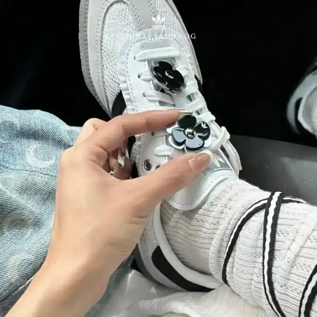 White Adidas Samba OG shoes with grey suede, customized with thick black and white striped laces and black floral accessories for a unique style.