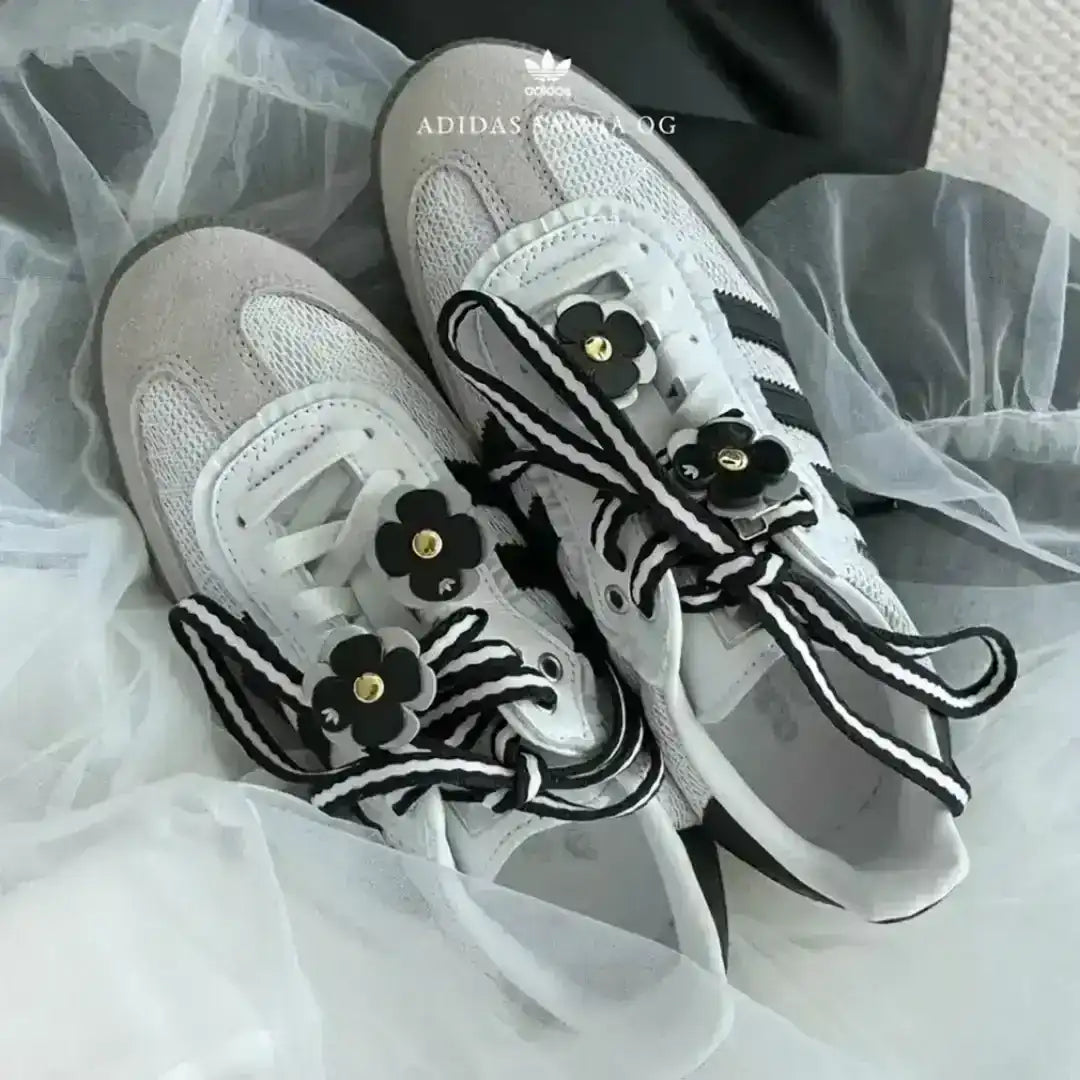 White Adidas Samba OG kicks stylized with custom details, including thick black and white striped laces and black floral patches with gold metal accents.