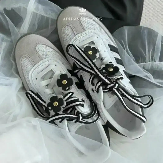 White Adidas Samba OG kicks stylized with custom details, including thick black and white striped laces and black floral patches with gold metal accents.
