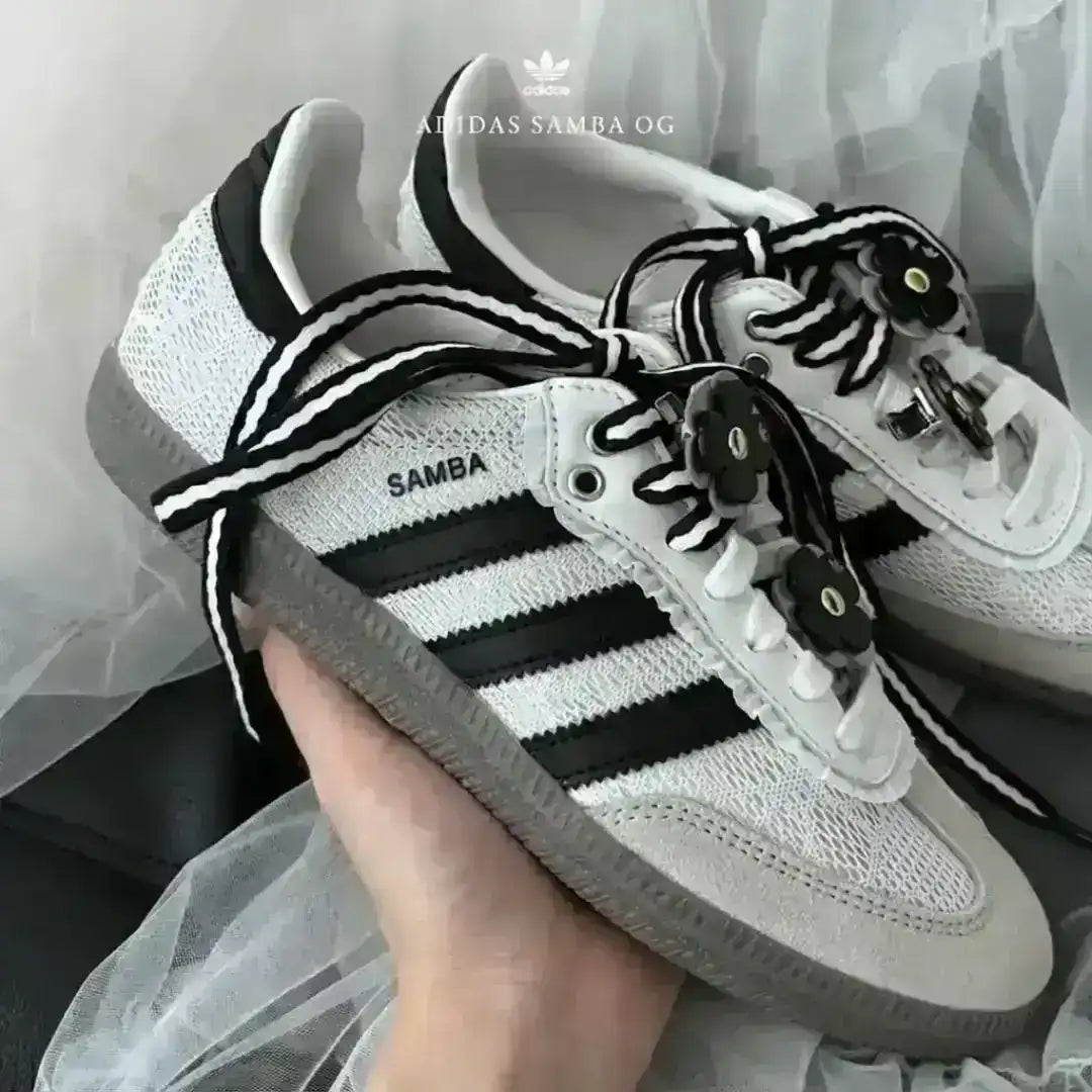 Adidas Samba OG white sneakers resting on a sheer white fabric, customized with black and white striped laces and removable flower charms.