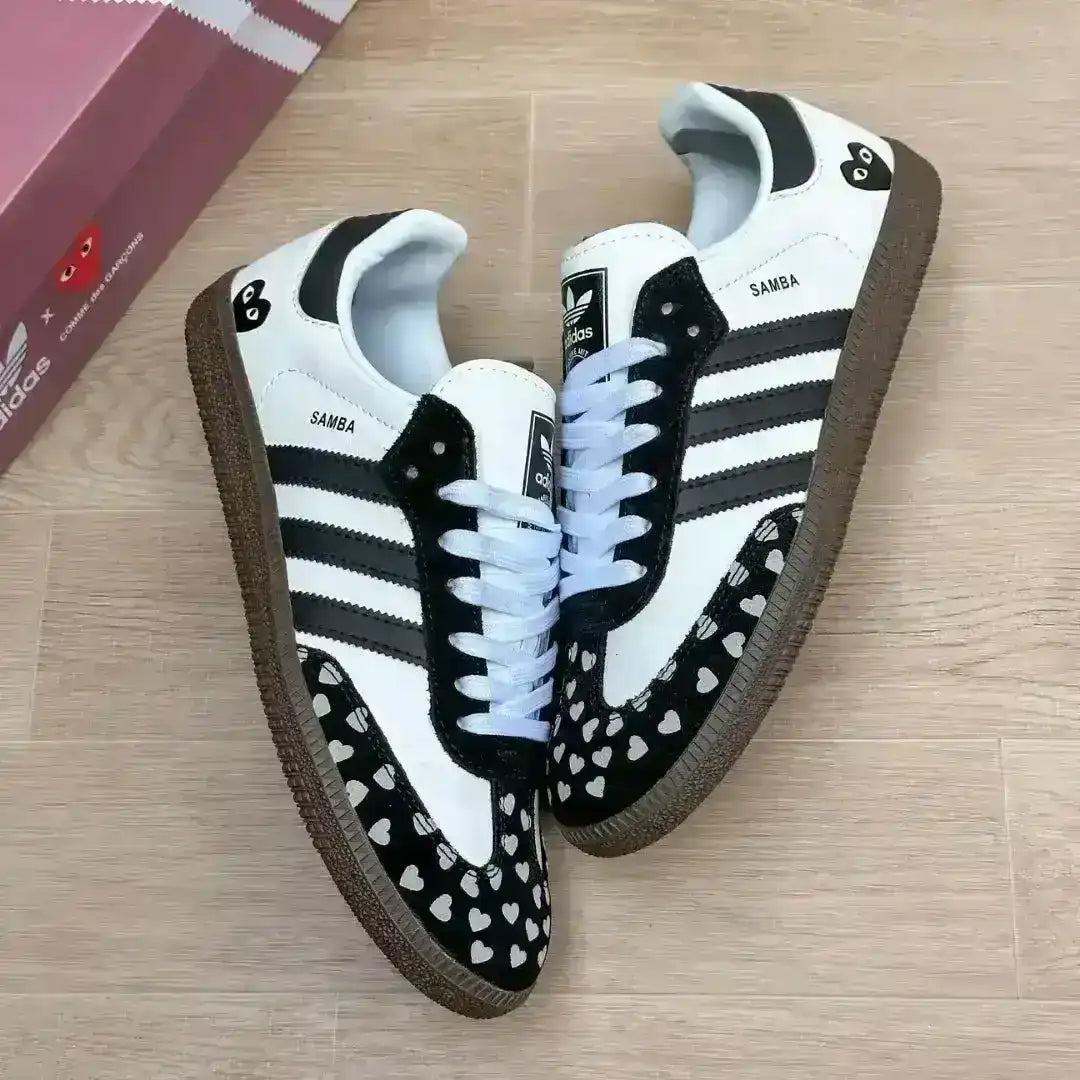 Trendy Adidas Samba footwear customized with a black and white theme, highlighting the "SAMBA" gold lettering and a distinctive heart print design on the toe box.