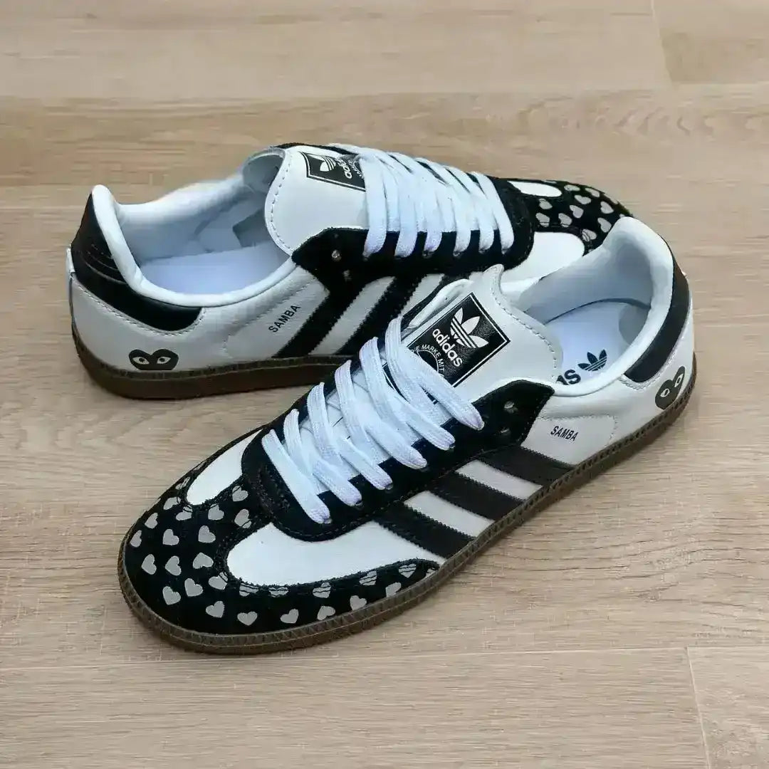 Pair of Adidas Samba trainers featuring a white leather upper, black heel tab with a heart icon, and a custom black suede toe section covered in small white hearts.